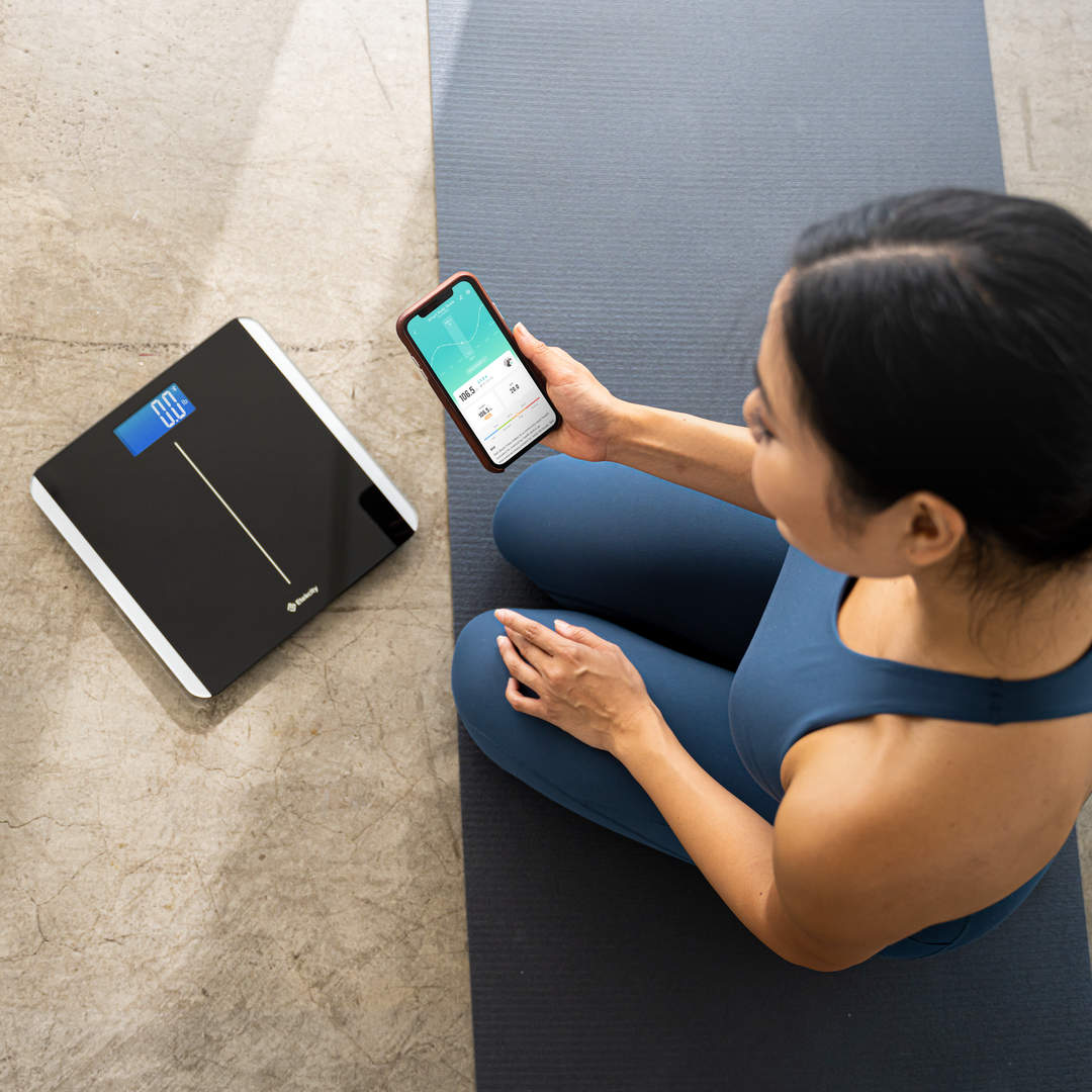 Woman exercising next to the Etekcity Smart Body Weight Scale with VeSync app on smartphone