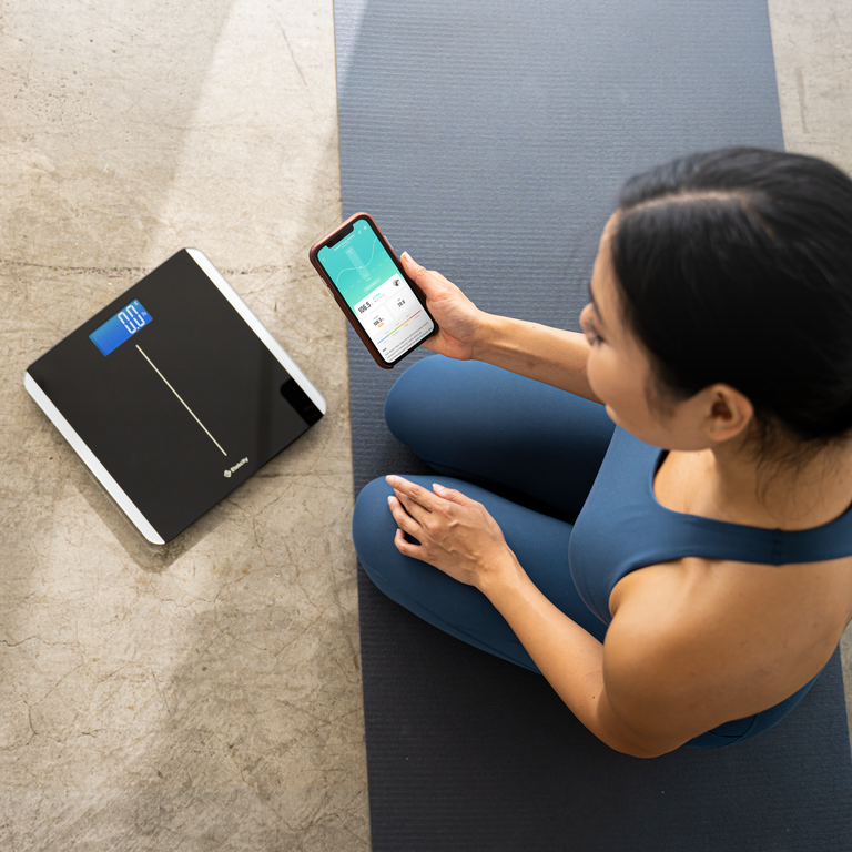 Woman exercising next to the Etekcity Smart Body Weight Scale with VeSync app on smartphone 