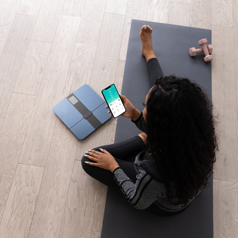 A woman stretching while looking at VeSync next to the Etekcity HR Smart Fitness Scale 